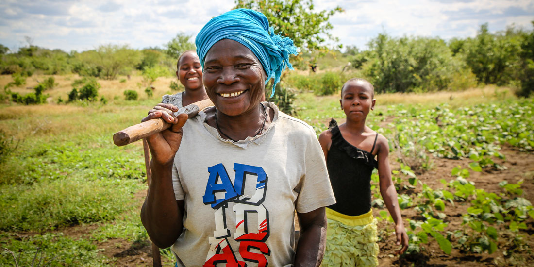 a Kenyan woman smiling with a stick over her shoulder and two younger women walking behind her in a farming plot