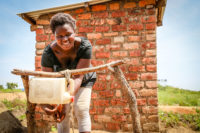 African woman smiling and washing her hands using a bucket tied to a wooden frame