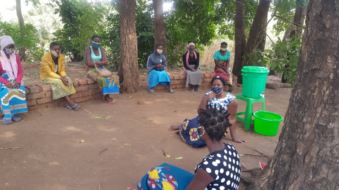 A group of women in Malawi sit together at a distance wearing face masks