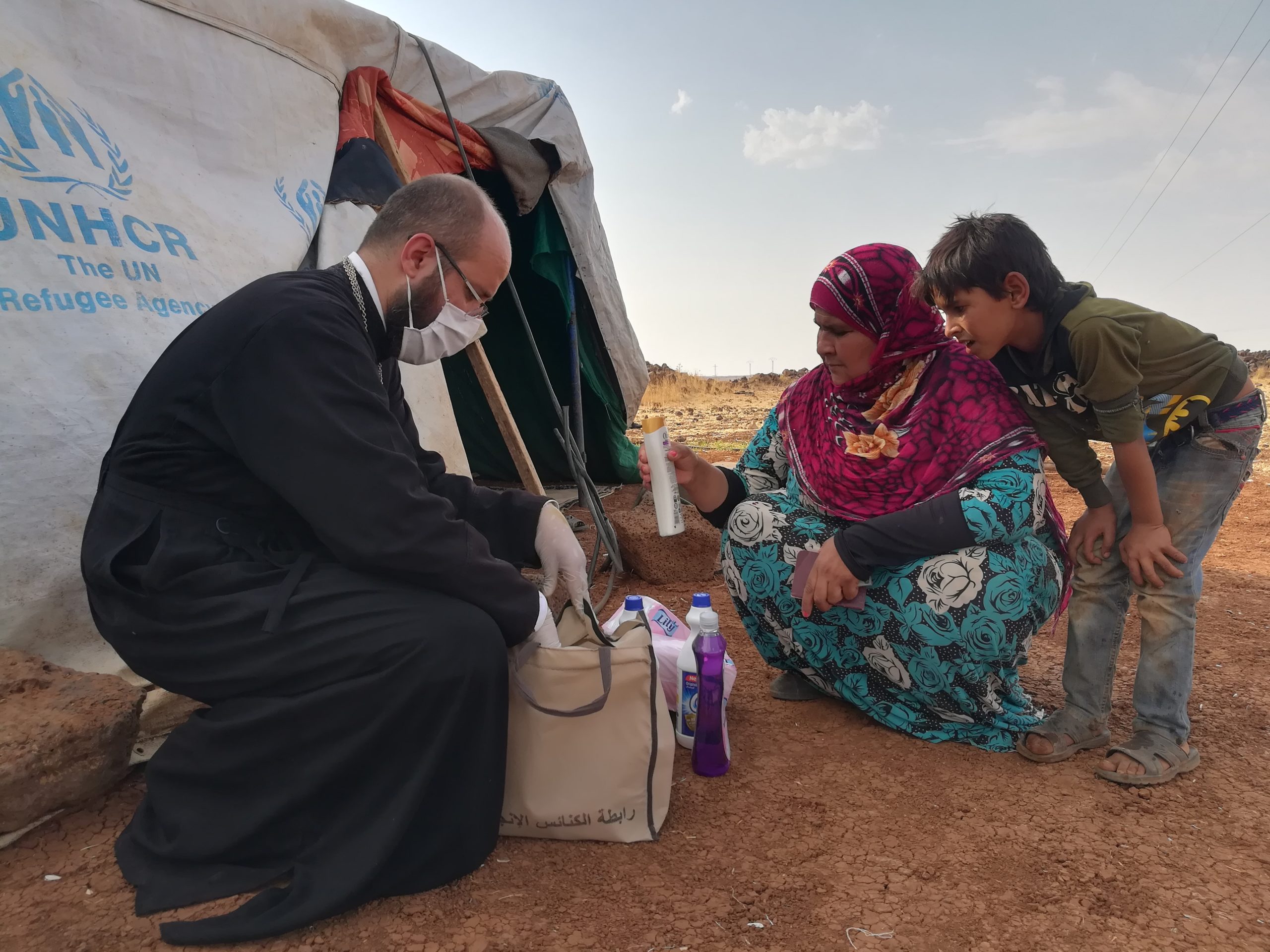 The United Nations High Commissioner for Refugees staff member with a medical face masks provides hygiene supplies to a mother and son