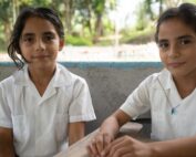 Two Honduran students in white school uniforms sit at wooden desks in an outdoor classroom area surrounded by trees.
