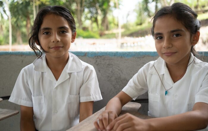 Two Honduran students in white school uniforms sit at wooden desks in an outdoor classroom area surrounded by trees.