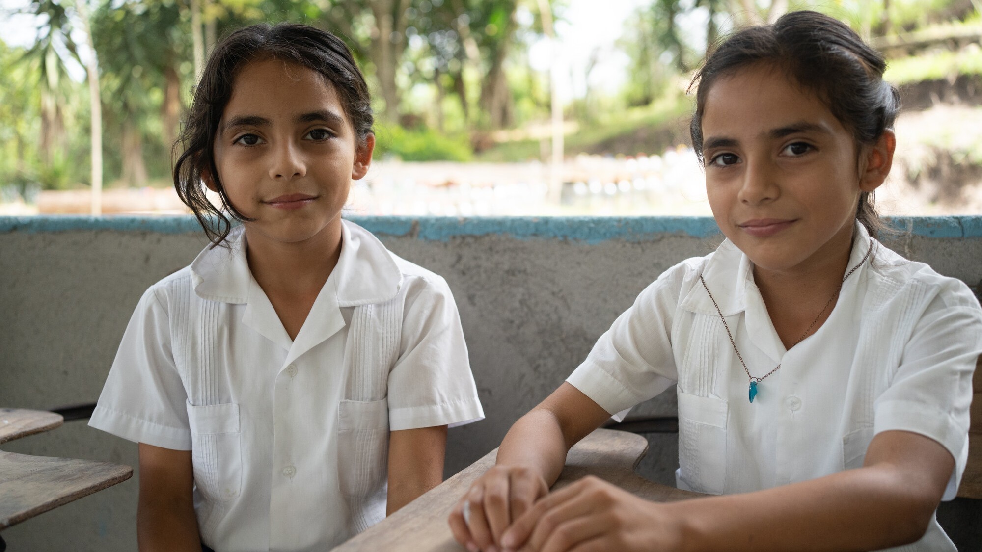 Two Honduran students in white school uniforms sit at wooden desks in an outdoor classroom area surrounded by trees.