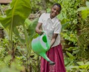 A girl in a school uniform waters plants in a lush garden with a bright green watering can.