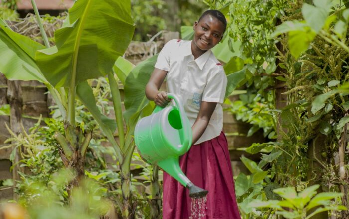 A girl in a school uniform waters plants in a lush garden with a bright green watering can.
