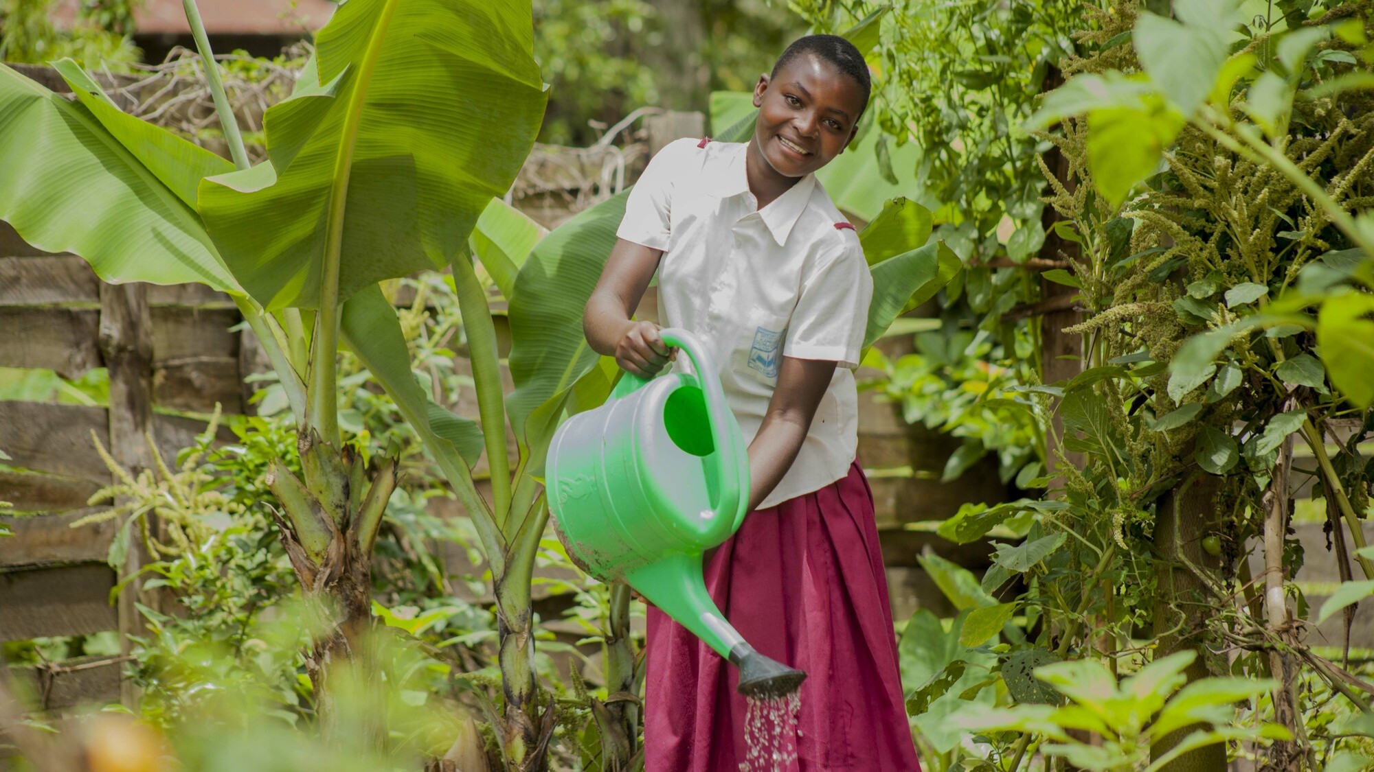 A girl in a school uniform waters plants in a lush garden with a bright green watering can.