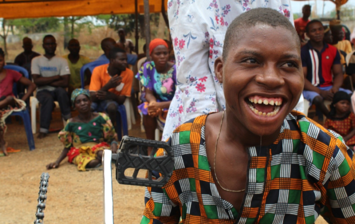 Person using a tricycle wheelchair outdoors with a group of seated people in the background.