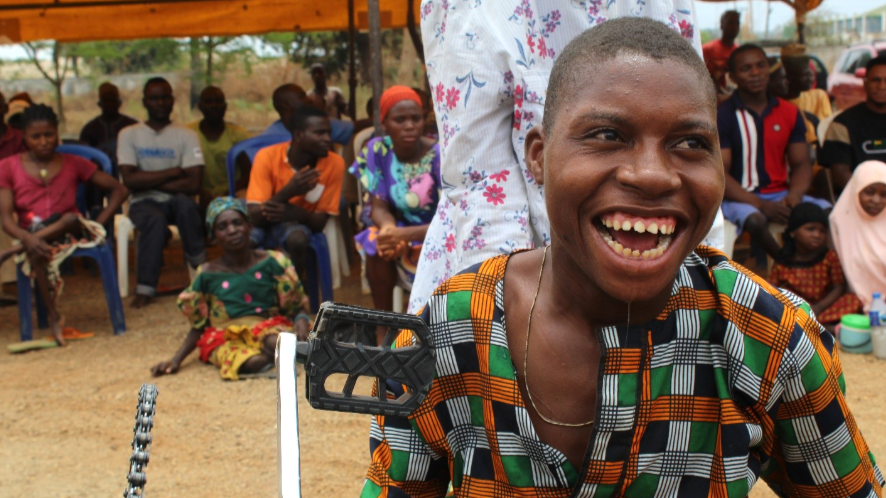 Person using a tricycle wheelchair outdoors with a group of seated people in the background.